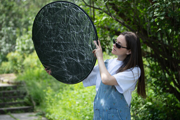 A young woman wearing sunglasses and casual attire is holding a round reflector while preparing for a photoshoot in a vibrant garden setting. Bright sunlight enhances the natural colors