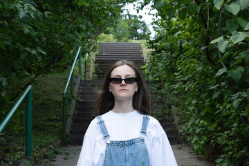 A young individual wearing a white shirt and denim overalls confidently ascends steps in a lush green area. Sunlight filters through the trees, creating a vibrant atmosphere
