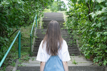 A young woman ascends stone steps bordered by lush greenery in a serene park. Sunlight filters through the leaves, creating a peaceful atmosphere