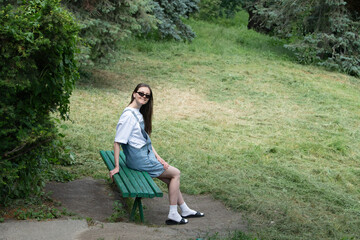 A young woman with long hair is seated on a green wooden bench in a park. She wears a casual outfit and sunglasses while relaxing in the bright afternoon sunshine surrounded by greenery