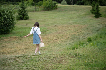 A young woman wearing denim overalls strolls through a lush green meadow, holding a bag. The peaceful afternoon atmosphere is filled with sunlight and tranquility
