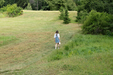 A girl strolls through a lush, green field surrounded by trees under clear skies. She carries a small basket, enjoying the peaceful rural atmosphere