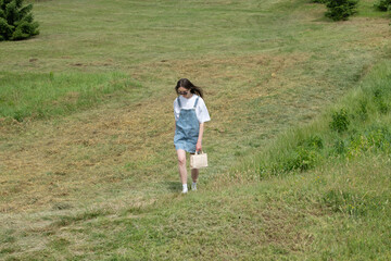 A girl strolls down a gentle slope covered in lush green grass. She wears a casual outfit and carries a small bag, enjoying a bright and pleasant day in the countryside