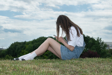 A young woman wearing a casual outfit sits on the grass, absorbed in her smartphone, surrounded by green trees and a cloudy sky, enjoying a peaceful moment outdoors