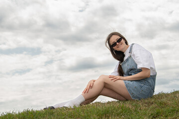 A young woman sits comfortably on a grassy hill, wearing denim overalls and sunglasses. The cloudy...