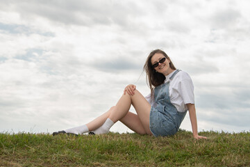 A young woman sits casually on a grassy hill wearing denim overalls and sunglasses. She smiles while enjoying the fresh air under a partly cloudy sky during a leisurely afternoon