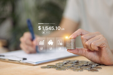 A hand holds a stack of coins in front of a digital financial dashboard while another hand writes, illustrating tracking savings and financial goals.