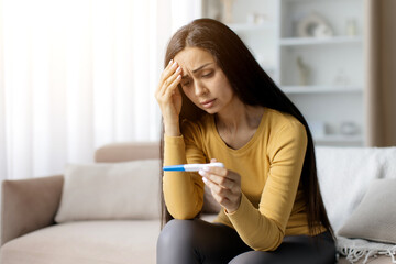 Worried young woman sitting on sofa, holding positive pregnancy test with hand on forehead,...