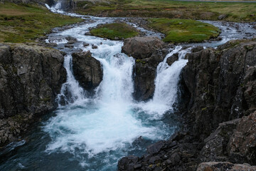 Powerful waterfall cascading through a rocky gorge in the rugged Icelandic landscape, with a winding river leading into the rapids and green banks