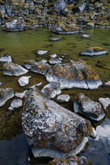 Close-up of moss-covered rocks in clear shallow water with a rocky shoreline. The natural textures and earthy tones create a serene, untouched landscape in a remote wilderness
