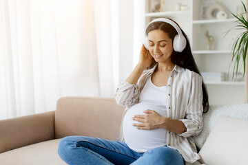 Beautiful pregnant woman in wireless headphones listening to music and touching her tummy while sitting on couch at home, smiling lady relaxing in living room, copy space