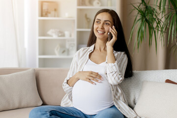 Obraz premium Pregnant woman talking on mobile phone while relaxing on a sofa at home, smiling joyfully with hand on belly, enjoying positive conversation in a bright living room, copy space