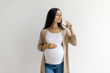 Young pregnant woman enjoying glass of water at home and tenderly embracing belly, standing near...
