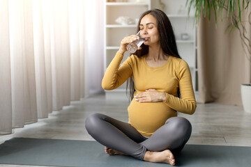 Pregnant woman sitting crosslegged on yoga mat, drinking water with eyes closed, enjoying hydration...