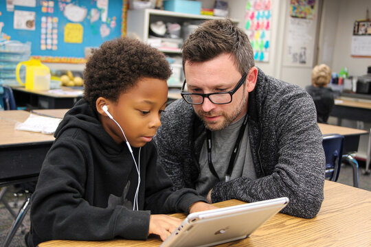 Teacher and student in elementary classroom use an iPad together, leaning on desk while working. Boy in black hoodie engaged in interactive learning and support.

