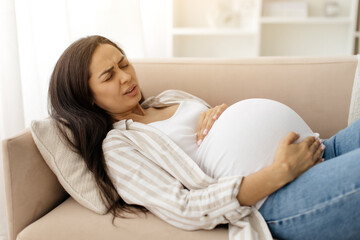 Young pregnant woman lying on sofa with pained expression, holding her belly during a contraction,...