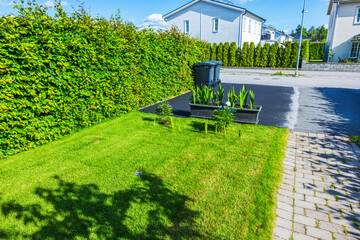 View of sunny front yard with green lawn, flowerbeds and driveway leading to two parking spots beside residential street. Sweden.