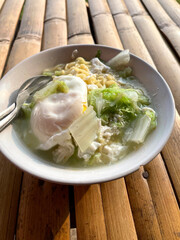 Hand movements holding spoons and forks, stirring a bowl of delicious steaming instant noodles, complemented by fried eggs and chopped green vegetables such as cabbage. The bowl is placed on a bamboo 