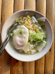 Hand movements holding spoons and forks, stirring a bowl of delicious steaming instant noodles, complemented by fried eggs and chopped green vegetables such as cabbage. The bowl is placed on a bamboo 