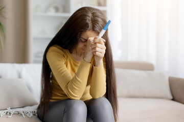Depressed woman holding pregnancy test in both hands and crying, head lowered in emotional moment,...