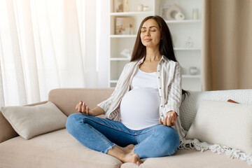 Beautiful young pregnant woman practicing yoga at home, sitting in lotus position on couch in...