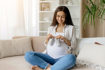 Pregnant Woman Preparing For Motherhood, Holding Small Baby Shoes Near Belly, Happy Awaiting Lady Sitting On Couch And Smiling, Enjoying Maternity, Copy Space