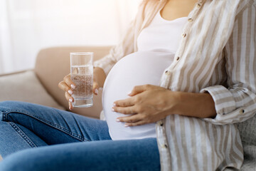 Closeup of pregnant woman touching belly and holding clear glass of sparkling water, taking healthy...