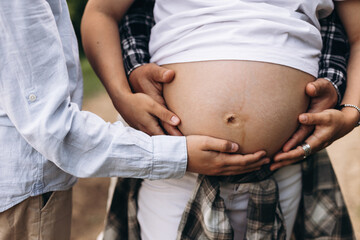 Close-up of a pregnant woman's belly with multiple hands gently embracing it. The woman is wearing...