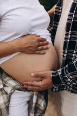 Close-up of a pregnant woman's belly with multiple hands gently embracing it. The woman is wearing a white shirt and plaid shirt tied around her waist. The photo conveys love, anticipation, and family