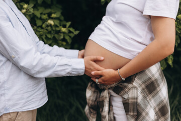 Close-up of a pregnant woman's belly with multiple hands gently embracing it. The woman is wearing a white shirt and plaid shirt tied around her waist. The photo conveys love, anticipation, and family
