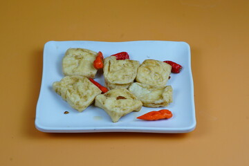 A close up of several pieces of fried tofu, a common Indonesian snack, served on a white plate with red chilies, against an orange background.
