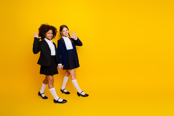 Two preteen schoolgirls in uniforms smiling and waving hands on a yellow background, celebrating friendship and education
