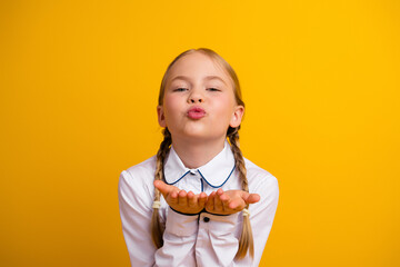 Happy schoolgirl in a uniform sending a playful kiss gesture against a vibrant yellow background in...