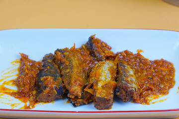 A rectangular white plate holds several pieces of fried mackarel tuna fish in sambal balado or a rich, spicy chilli sauce, set against a plain yellow background.