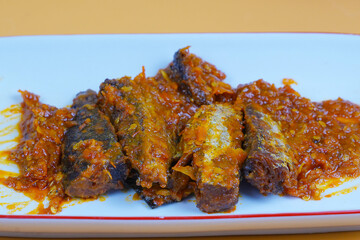 A rectangular white plate holds several pieces of fried mackarel tuna fish in sambal balado or a rich, spicy chilli sauce, set against a plain yellow background.