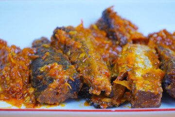 A rectangular white plate holds several pieces of fried mackarel tuna fish in sambal balado or a rich, spicy chilli sauce, set against a plain yellow background.