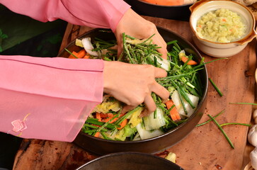 A woman wearing a pink hanbok is sitting and kneading cabbage in a large bowl to make kimchi....