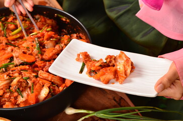 A person in a Hanbok serves freshly made, vibrant kimchi from a large pan onto a white rectangular dish, showcasing the bright red spice and fresh green vegetables.