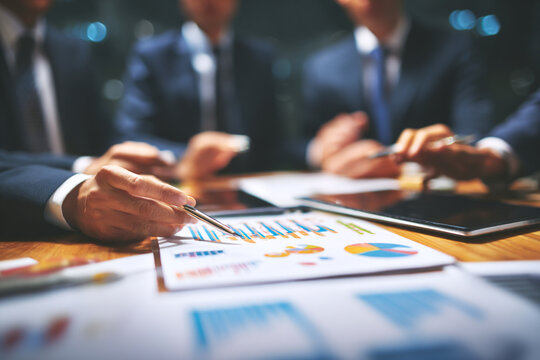 Group of business professionals in suits gathered around office table reviewing charts, graphs, and digital tablets. Corporate teamwork, financial analysis, strategic planning, and modern business col