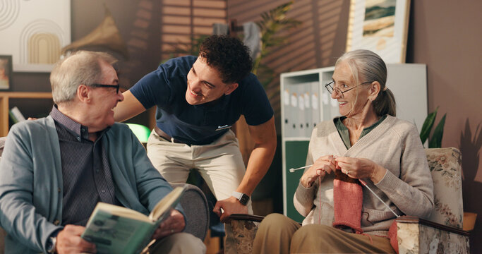 Living room, caregiver and talk with old couple, hobby and senior people with smile in nursing home. Knitting, elderly woman and man with book in lounge, nurse and conversation with patient in house - Powered by Adobe