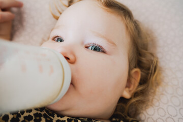 A young child with curly hair is drinking from a bottle propped up comfortably on a soft surface at...