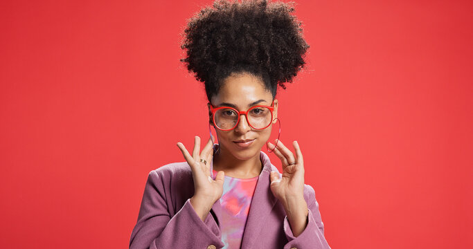 Portrait, African woman and fashion in studio with earrings, glasses and trendy clothes. Proud, female person and positive attitude with jewellery, stylish outfit and accessories on red background - Powered by Adobe