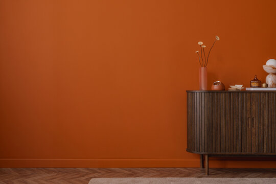 Minimalist interior with decorative objects and a pink ribbed vase on a wooden cabinet against a plain burnt orange wall. Calm, modern composition. Copy space.