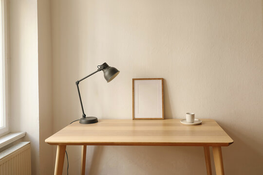 Elegant Scandinavian interior, living room. Blank thin wooden picture frame mockup on table near window. Modern lamp. Cup of coffee, tea. Trendy working space, home office. Beige wall background