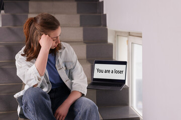 Cyber bullying. Upset teenage girl and laptop with text You Loser on stairs indoors