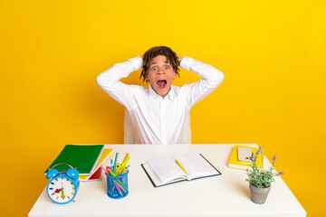 Excited schoolboy with hands on head sitting at a study desk filled with school supplies against...