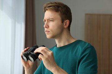 Paranoia. Worried man with binoculars near window indoors