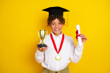 Smiling schoolboy in graduation attire holding a trophy and diploma, celebrating academic success...