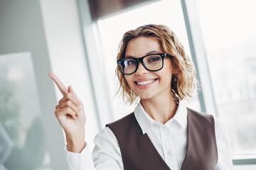 Smiling businesswoman in formal attire pointing upwards in a modern office environment with large...