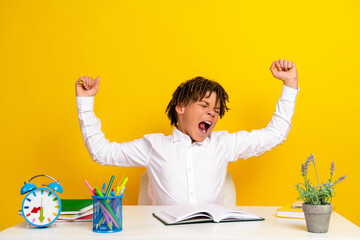 Young boy in white shirt stretching during study at desk with school supplies against bright yellow...